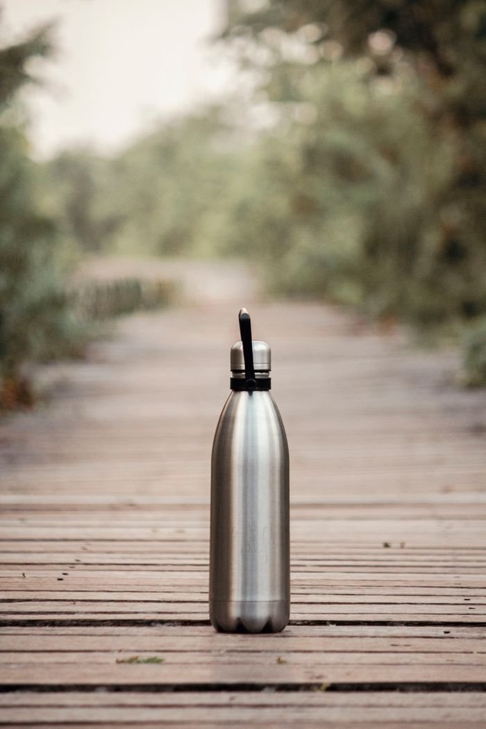 A stainless steel water bottle on a wooden path in an outdoor setting, surrounded by greenery.