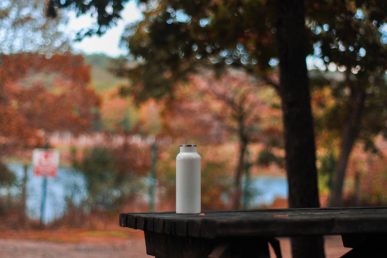 A stainless steel water bottle sits on a picnic table in an autumn park setting with colorful foliage.