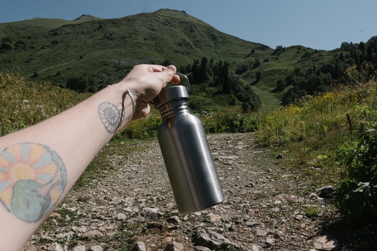 A tattooed arm holding a stainless steel bottle against a scenic mountain backdrop.